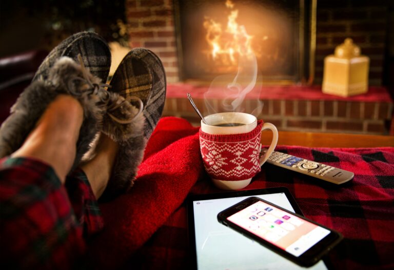 Person with feet up on a table in front of a fireplace with a mug of hot coco next to a tablet and smart phone