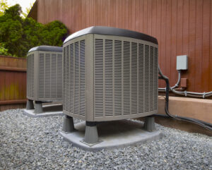 A low-angle outdoor photo of two residential air conditioning units. The units are gray with black tops and are placed on separate concrete pads surrounded by light-colored gravel. They are positioned against a dark brown wooden fence or wall. A few electrical lines and a utility box are visible on the wall.