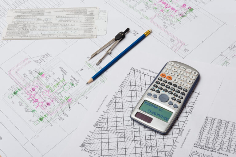 n overhead photo shows a variety of engineering or architectural documents laid out on a table. The papers are covered with various schematics, charts, and blueprints. A calculator, a blue pencil, and a drawing compass are resting on top of the documents.