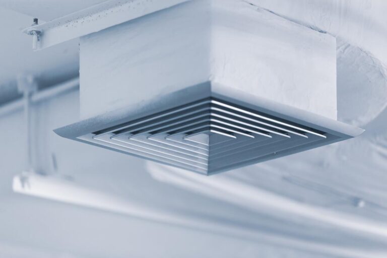 A close-up, low-angle photo of a modern, square-shaped air conditioning vent on a white ceiling. The vent has multiple horizontal louvers and is connected to the ceiling by a white duct. The overall color of the image is a cool, light blue, and the image is sharply focused on the vent itself.
