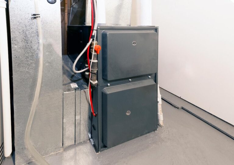 A low-angle photo shows a modern residential furnace unit. The unit is a dark gray color with two access panels on the front. It is connected to a larger, silver metal duct on the left. The units are sitting on a light gray concrete floor against a plain white wall. A few pipes and wires are visible on the left side of the gray unit.
