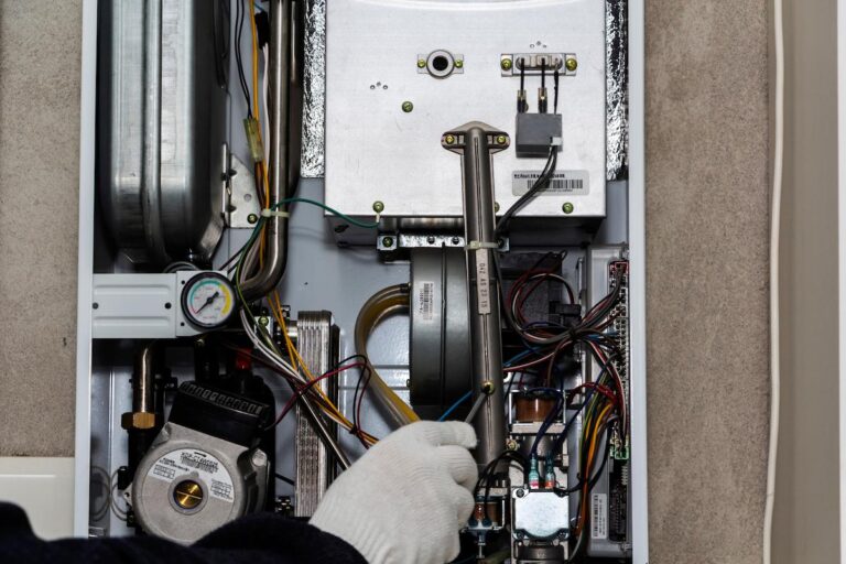 A close-up photo shows the interior components of a furnace or boiler. A hand wearing a white glove is reaching in to perform maintenance or a repair. The unit is a jumble of wires, pipes, and various metal parts, including a pressure gauge on the left side. The image is cropped to show only the internal workings and the gloved hand.