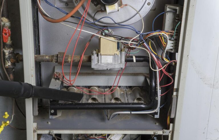 A close-up photo shows the interior of an open furnace or heating unit. A black vacuum hose is being used to clean dust and debris from the burners at the bottom of the unit. The inside is a complex arrangement of metal pipes, burners, and a jumble of multi-colored wires. The image is cropped to show the internal parts being serviced.