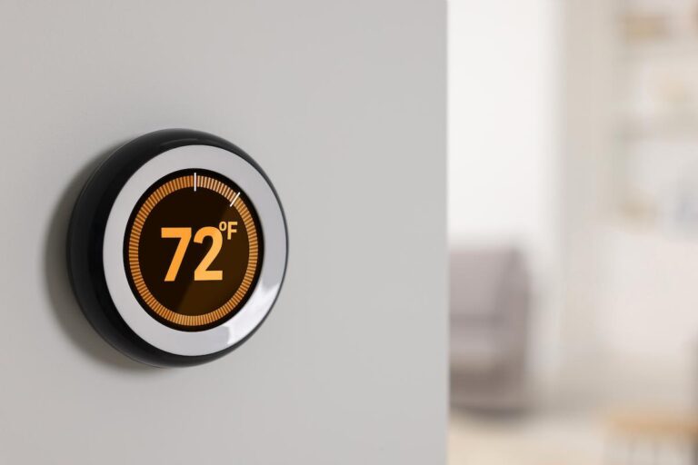 A close-up photo of a modern, round smart thermostat mounted on a light-colored wall. The digital display is glowing with an orange light, showing the temperature set to "72°F." The background is a blurry, sunlit room, with a couch visible in the distance.