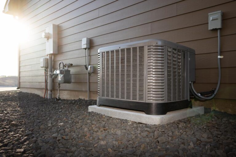 A professional photo of a residential air conditioning unit on a concrete pad, sitting on a bed of dark gray gravel. The unit is gray with a black base. It is placed against a brown-colored house with horizontal siding. To the left of the unit, various utility boxes, pipes, and electrical lines are visible on the side of the house. The sun is shining brightly from the top left corner, creating a lens flare.