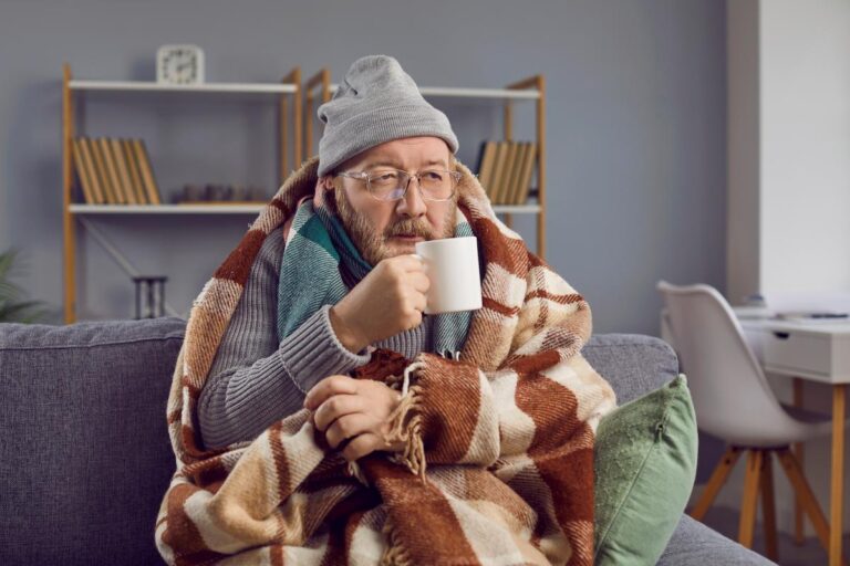 A medium shot of a man with fair skin and a beard, wearing a gray beanie and glasses. He is sitting on a gray couch, bundled up in a brown and white patterned blanket and a blue scarf, and holding a white mug to his mouth. He has a concerned expression on his face, suggesting he is cold or sick. The background is a blurry living room with a bookshelf and a white chair.