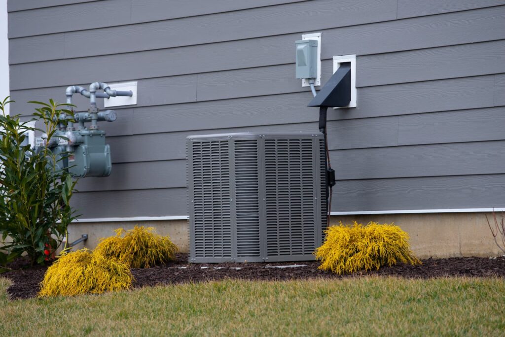 A low-angle photo of a residential air conditioning unit positioned against a house with gray horizontal siding. A gas meter and associated piping are visible on the left. The ground is covered with mulch, green grass, and three bright yellow-green shrubs. The sky is overcast and gray.