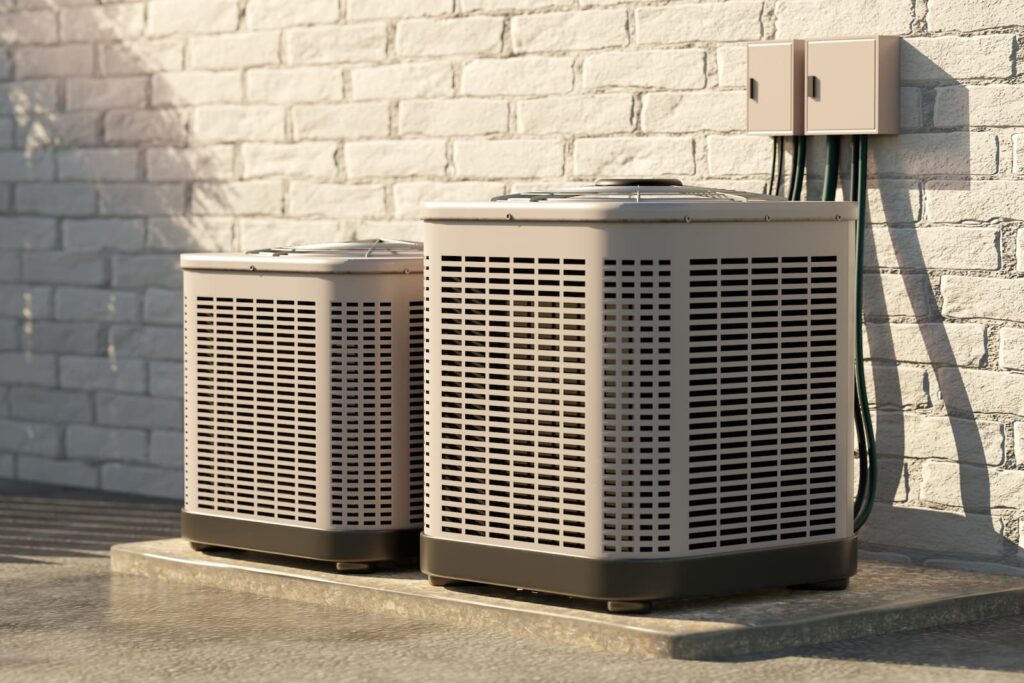 A low-angle photo of two identical beige and black air conditioning units sitting on a concrete pad outside a building. The building wall behind the units is made of light-colored brick. Electrical lines run from a small beige utility box on the wall to the unit in the foreground. The sunlight hits the scene from the left, casting sharp shadows.
